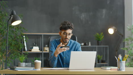 Arabic-Young-Man-In-Eyeglasses-Sitting-At-Desk,-Holding-Credit-Card-And-Shopping-Online-On-Laptop-Computer