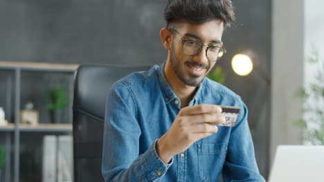 Close-Up-View-Of-Arabic-Young-Man-In-Eyeglasses-Sitting-At-Desk,-Holding-Credit-Card-And-Shopping-Online-On-Laptop-Computer