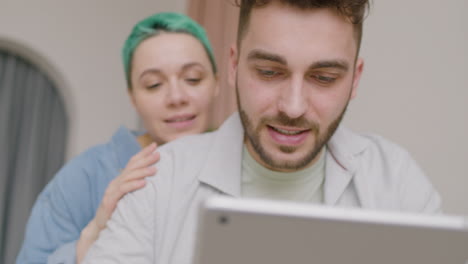 Close-Up-View-Of-Young-Man-Using-A-Tablet-While-His-Girlfriend-Hugging-Him-From-His-Back
