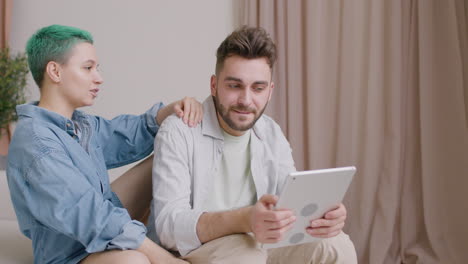 Man-Holding-A-Tablet-While-Talking-With-His-Girlfriend-Sitting-On-Sofa-At-Home