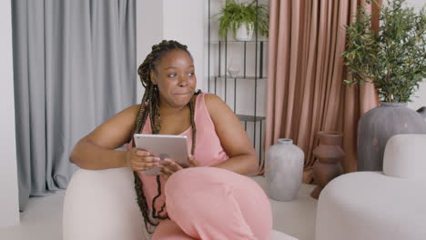 Woman-With-Braids-Sitting-On-A-Big-Sofa-While-Using-A-Tablet,-Then-Looks-At-A-Side