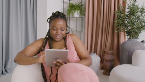 Woman-With-Braids-Lying-On-A-Big-Sofa-While-Smiling-And-Using-A-Tablet