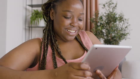 Close-Up-View-Of-Woman-With-Braids-Lying-On-A-Big-Sofa-While-Smiling-And-Using-A-Tablet