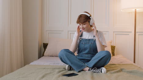 Front-View-Of-Girl-Sitting-On-Bed-Wearing-Headphones-And-Playing-Music-From-Her-Smartphone