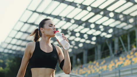 Young-Jogger-Woman-Training-In-The-Stadium-And-Drinking-Water-From-A-Bottle-In-A-Summer-Morning