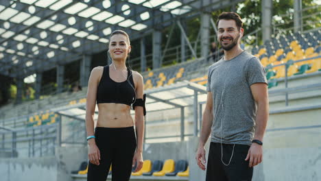Young-Jogger-Couple-Looking-At-Each-Other-And-Then-Smiling-To-The-Camera-While-They-Are-Training-In-The-Stadium-On-Summer-Day