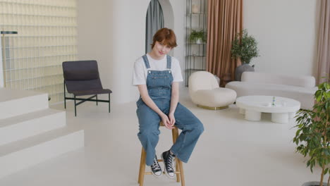 Girl-In-Denim-Jumpsuit-Sitting-On-Banquette-And-Looking-At-Camera-And-Smiling-In-The-Middle-Of-The-Living-Room-Of-A-Modern-House