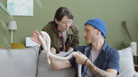 Couple-Holding-Pet-Snakes-And-Talking-Together-While-The-Man-Is-Sitting-And-Woman-Is-Leaning-On-A-Comfortable-Sofa-At-Home-1