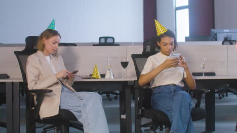 Two-Female-Colleagues-Sitting-In-Office-Chairs-And-Using-Mobile-Phone-During-A-Party-At-The-Office
