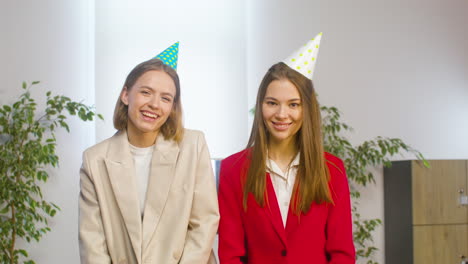 Two-Happy-Female-Colleagues-With-Party-Hat-Hugging-And-Looking-At-Camera-Ath-The-Office-Party