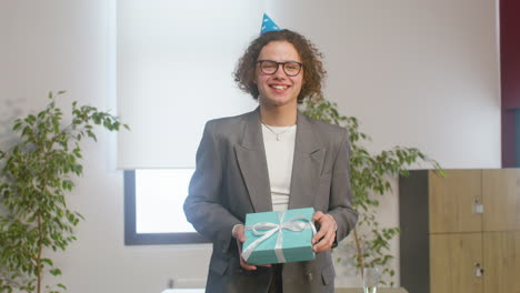 Portrait-Of-A-Happy-Curly-Boy-With-Party-Hat-Holding-A-Gift-Box-And-Looking-At-The-Camera-During-A-Office-Party