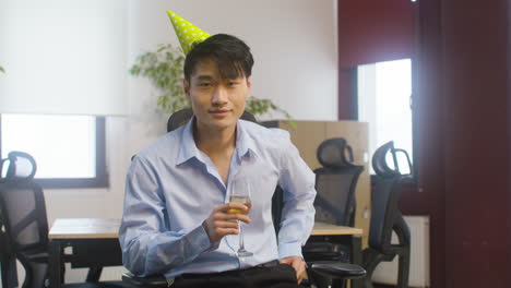 Portrait-Of-A-Confident-Man-In-Formal-Outfit-And-Party-Hat-Holding-A-Champagne-Glass-And-Looking-At-Camera-While-Sitting-At-The-Office
