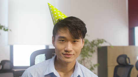 Portrait-Of-A-Smiling-Man-Wearing-A-Party-Hat-And-Looking-At-Camera-While-Confetti-Falling-At-The-Office