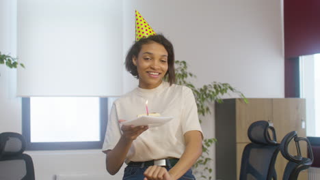 Portrait-Of-A-Happy-American-Girl-Holding-A-Slice-Of-Birthday-Cake-On-A-Plate-While-Looking-At-The-Camera-At-The-Office-Party