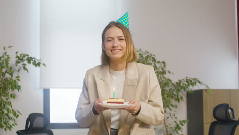 Portrait-Of-A-Happy-Girl-Holding-A-Slice-Of-Birthday-Cake-On-A-Plate-And-Blowing-Candle-While-Looking-At-The-Camera-At-The-Office-Party