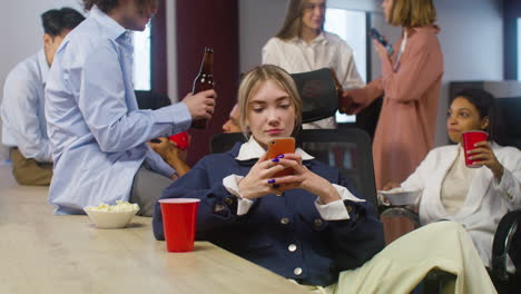 Blondes-Mädchen-Sitzt-Und-Benutzt-Handy,-Während-Ihre-Kollegen-Auf-Der-Büroparty-Reden-Und-Getränke-Anstoßen