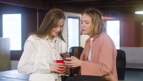 Two-Female-Colleagues-Holding-Drinks-And-Looking-Something-On-The-Mobile-Phone-During-A-Party-In-The-Office