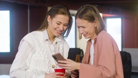 Two-Female-Colleagues-Holding-Drinks-And-Looking-Something-On-The-Mobile-Phone-During-A-Party-In-The-Office-1