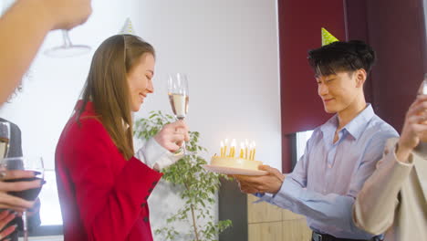 Happy-Woman-Blowing-Out-Candles-On-Cake-And-Drinking-With-Her-Multiethnic-Colleagues-At-The-Office-Party