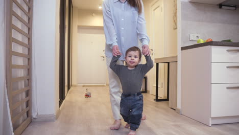 Mother-Helping-Her-Baby-To-Take-His-First-Steps-From-The-Corridor-To-The-Living-Room-At-Home