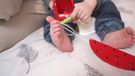 Camera-Focuses-On-The-Bare-Feet-Of-A-Baby-While-He-Is-Playing-With-Toys-On-The-Bed-With-His-Mother