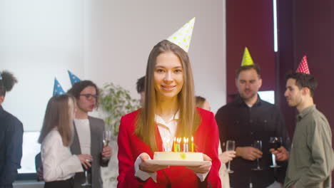 Portrait-Of-A-Beautiful-Woman-Holding-A-Birthday-Cake,-Closing-Eyes,-Making-A-Wish-And-Blowing-Out-Candles-While-Looking-At-Camera-During-A-Party-In-The-Office