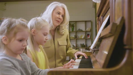 Two-Little-Girls-Playing-Old-Piano-At-Home-While-Their-Grandmother-Sitting-Next-To-Them