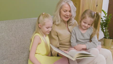 Grandmother-Sitting-On-Sofa-With-Her-Two-Granddaughters-And-Reading-Them-Fairy-Tales