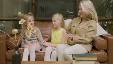 Happy-Grandmother-Sitting-On-Sofa-And-Hugging-Her-Two-Little-Granddaughters-At-Home-1