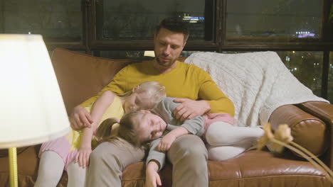 Happy-Father-Sitting-On-Sofa-With-His-Two-Sleepy-Little-Girls-Who-Leaning-Their-Heads-On-His-Lap