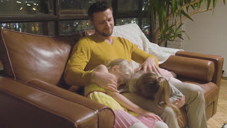 Happy-Father-Sitting-On-Sofa-With-His-Two-Little-Girls-Who-Sleeping-And-Leaning-Their-Heads-On-His-Lap