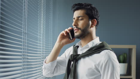 Nahaufnahme-Eines-Arabischen-Geschäftsmannes,-Der-In-Seinem-Büro-Am-Fenster-Steht-Und-Telefoniert-Und-Dann-Aus-Dem-Fenster-Schaut