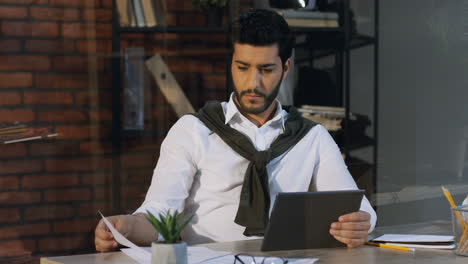 Junger-Arabischer-Geschäftsmann,-Der-In-Seinem-Büro-Am-Schreibtisch-Sitzt-Und-Arbeitet,-Während-Er-Daten-Des-Dokuments-Und-Auf-Dem-Tablet-Vergleicht