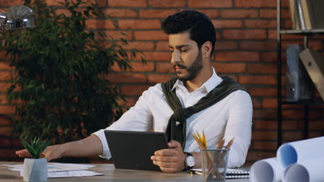 Young-Arabian-Architect-Sitting-At-Desk-In-His-Office-And-Working-With-Some-Documents-And-Tablet