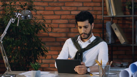 Junger-Arabischer-Geschäftsmann-Scrollt-Und-Klebt-Auf-Dem-Tablet,-Während-Er-Im-Modernen-Büro-Sitzt