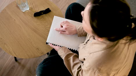 Blind-Woman-Reading-A-Braille-Book-While-Sitting-On-Sofa-At-Home