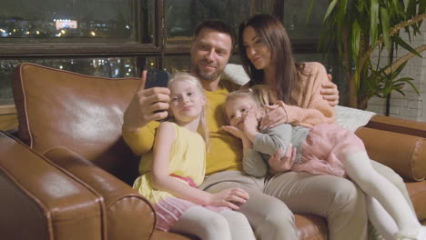 Happy-Parents-And-Two-Little-Girls-Taking-Selfie-Photo-While-Sitting-On-Sofa-At-Home