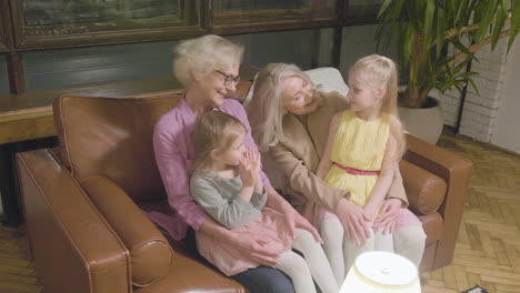 Two-Grandmothers-Talking-And-Spending-Time-With-Their-Two-Granddaughter-While-Sitting-On-Sofa-At-Home