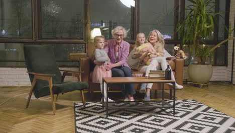 Zwei-Kleine-Mädchen-Rennen-Und-Umarmen-Ihre-Glücklichen-Großmütter,-Die-Zu-Hause-Auf-Dem-Sofa-Im-Wohnzimmer-Sitzen