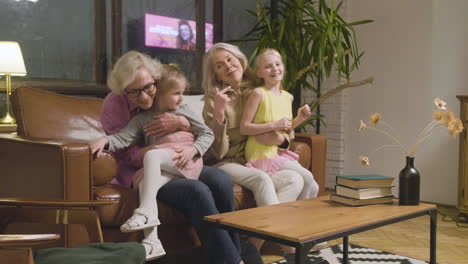 Two-Little-Girls-Running-And-Hugging-Their-Happy-Grandmothers-Who-Sitting-On-Sofa-In-The-Living-Room-At-Home-1