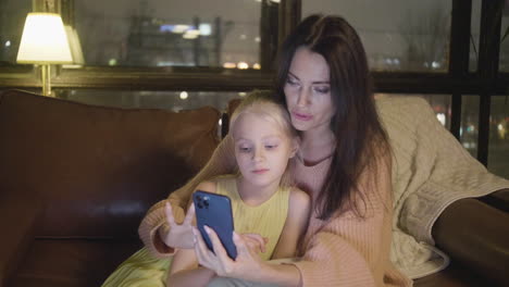 Woman-Watching-Something-On-Mobile-Phone-With-Her-Little-Daughter-While-Sitting-Together-On-Sofa-At-Home-At-Night