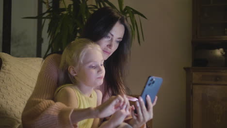 Woman-Watching-Something-On-Mobile-Phone-With-Her-Little-Daughter-While-Sitting-Together-On-Sofa-At-Home-At-Night-1