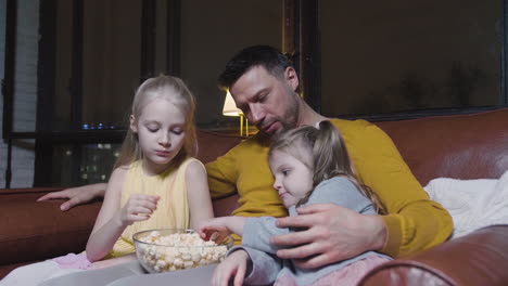 Dad-And-His-Two-Little-Daughters-Eating-Popcorn-And-Watching-A-Movie-While-Sitting-On-Sofa-At-Night-At-Home