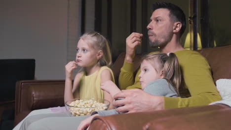 Vater-Und-Seine-Beiden-Kleinen-Töchter-Essen-Popcorn-Und-Schauen-Sich-Einen-Film-An,-Während-Sie-Nachts-Zu-Hause-Auf-Dem-Sofa-Sitzen-1