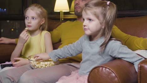Vater-Und-Seine-Beiden-Kleinen-Töchter-Essen-Popcorn-Und-Schauen-Sich-Einen-Film-An,-Während-Sie-Nachts-Zu-Hause-Auf-Dem-Sofa-Sitzen-2