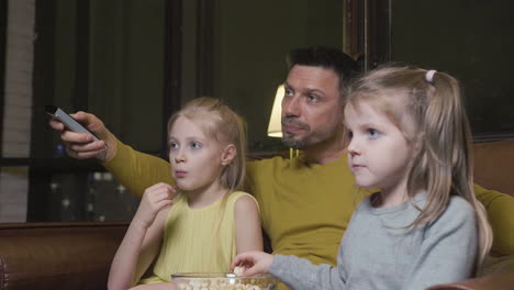 Dad-With-Tv-Remote-Control-And-His-Two-Little-Daughters-Eating-Popcorn-And-Watching-A-Movie-While-Sitting-On-Sofa-At-Night-At-Home