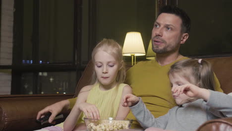 Dad-With-Tv-Remote-Control-And-His-Two-Little-Daughters-Eating-Popcorn-And-Watching-A-Movie-While-Sitting-On-Sofa-At-Night-At-Home-1