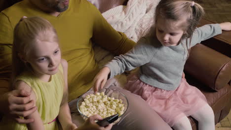 Dad-And-His-Two-Little-Daughters-Eating-Popcorn-And-Watching-A-Movie-While-Sitting-On-Sofa-At-Night-At-Home-3