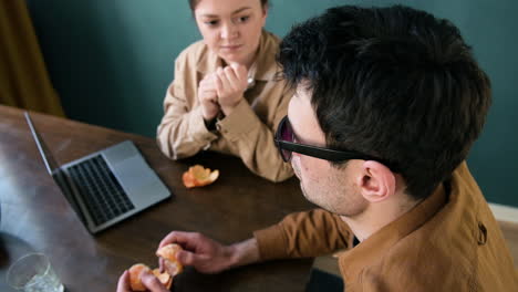 Over-The-Shoulder-Shot-Of-A-Woman-And-Blind-Man-With-Glasses-Eating-Tangerines-And-Talking-While-Sitting-At-Table-At-Home