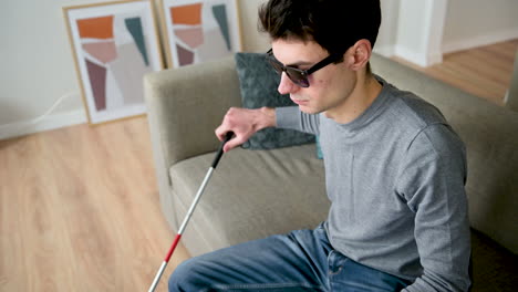 Blind-Man-Sitting-On-Sofa-At-Home,-Then-Wearing-Glasses,-Holding-Walking-Stick-And-Standing-Up